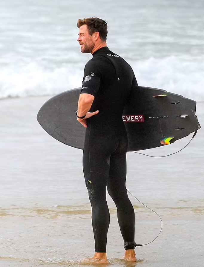 Киноактер Крис Хемсворт с доской для серфинга на берегу моря.