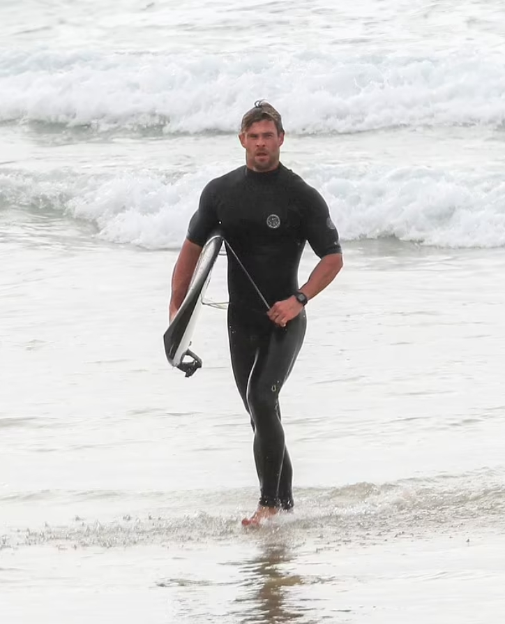 Киноактер Крис Хемсворт в плавательном костюме на фоне больших волн.