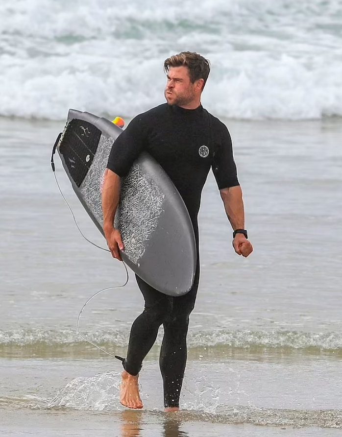 Киноактер Крис Хемсворт на берегу моря в черном плавательном костюме и с доской для серфинга.